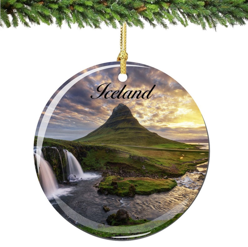 May include: Round glass Christmas ornament featuring a scenic view of a mountain range in Iceland with a waterfall in the foreground. The ornament has a gold string for hanging and the word "Iceland" printed on the top.