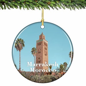 May include: A round, blue and white Christmas ornament featuring a photograph of a tall, red brick minaret with a green tiled roof. The text "Marrakesh Morocco" is printed in white on the ornament.