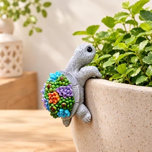 May include: A grey turtle figurine designed to hang on the rim of a planter. The turtle shell is decorated with a cluster of colourful miniature succulents in shades of blue, green, orange, and purple, featuring a textured skin and black eyes.