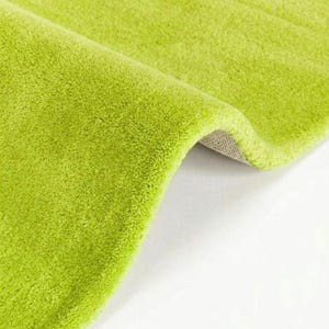 Pode incluir: Um tapete verde-limão brilhante com uma textura macia, felpuda e de pelo alto. O tapete está dobrado para revelar um forro de tecido entrelaçado de cor clara. A superfície do tapete apresenta uma cor sólida uniforme e um aspeto denso e felpudo.