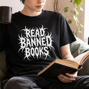 Puede incluir: Una camiseta negra de manga corta con texto gráfico blanco que dice READ BANNED BOOKS en una fuente estilizada, dentada e inspirada en el metal, centrada en el pecho. La camiseta tiene cuello redondo y un corte estándar de tela negra.