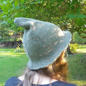 Puede incluir: Un gorro de punto verde con una corona puntiaguda y un ala ancha. El gorro está hecho con un hilo texturizado y tiene un efecto moteado sutil.