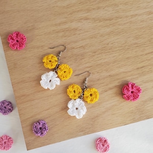 May include: Crocheted flower earrings in white, yellow, and pink. The earrings are displayed on a wooden surface with additional crocheted flowers in various colors.