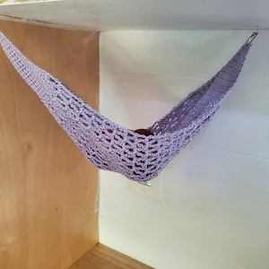 Puede incluir: Una hamaca de ganchillo lavanda suspendida del techo. La hamaca está hecha de un delicado patrón de tejido abierto. La hamaca cuelga en la esquina de una habitación, con paredes de madera y blancas visibles.