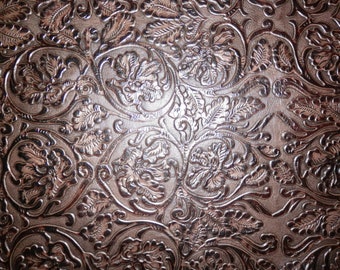 May include: Close-up of a brown leather surface with an embossed floral pattern. The design features stylized flowers, leaves, and swirling vines, creating a textured, decorative effect. The leather has a metallic sheen.