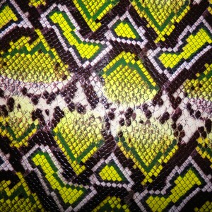 May include: Close-up of a textured pattern resembling snakeskin. The design features a repeating geometric pattern in shades of yellow, green, brown, and white, creating a striking visual effect. The pattern is likely used for fashion or decorative purposes.