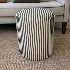 May include: A cylindrical ottoman featuring a vertical striped pattern in dark olive green and off-white fabric. The stool has a rounded top edge with piped detailing and a structured, upright form, providing a stable surface for seating or foot support.