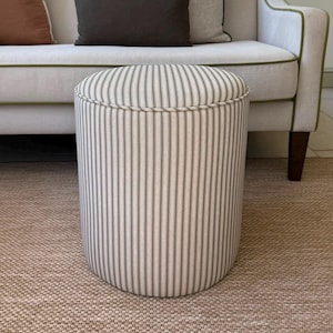 May include: A cylindrical ottoman upholstered in a cream-coloured fabric featuring thin, vertical dark green pinstripes. The top edge of the ottoman is finished with a matching fabric-covered piping detail that runs along the circular perimeter.