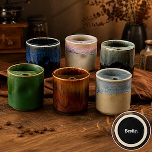 May include: Six cylindrical ceramic mugs with a unique notched rim and a small hole. The mugs feature varied reactive glazes in shades of deep blue, green, brown, beige, and pink. A circular inset shows the bottom of a mug with the text Bestie.