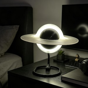 May include: A planet-shaped table lamp featuring a dark spherical center surrounded by a glowing, translucent ring structure. The lamp is supported by a black curved arm attached to a circular base, emitting a bright white light from the ring.
