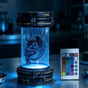 May include: A cylindrical lamp featuring a dark, textured base and top with yellow accents, containing a detailed grey alien creature figure inside a clear tube. The lamp emits a blue light and includes a remote control with multiple colour buttons.
