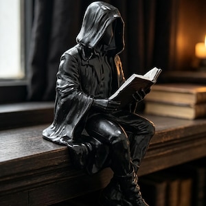 May include: A black figurine of a seated figure wearing a hooded cloak and reading an open book. The figure is depicted in a contemplative pose with the hood pulled over the head, obscuring the face, while holding the book with both hands.