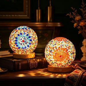 May include: Two spherical table lamps featuring intricate mosaic glass patterns in vibrant shades of blue, red, yellow, and white. Each lamp sits on a circular light-colored wooden base with a small power button, emitting a warm, soft glow.