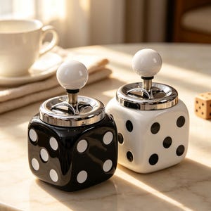 May include: Two square ceramic containers shaped like dice, one black with white dots and one white with black dots. Each container features a silver-toned metal lid with a central push-button mechanism topped by a small white spherical knob.