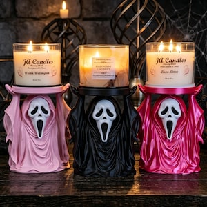 Peut inclure: Trois bougies en pot en verre à trois mèches, reposant sur des supports en forme de fantôme. Les supports sont rose clair, noir et rose vif, chacun arborant un masque blanc avec une expression à la bouche grande ouverte.