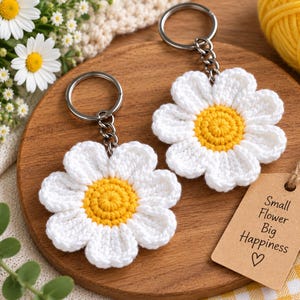 Puede incluir: Dos llaveros de margarita tejidos a ganchillo con pétalos blancos y centro amarillo, cada uno unido a una anilla de metal plateado. Incluye una etiqueta de papel marrón con el texto impreso: Small Flower Big Happiness y un corazón.