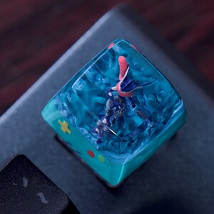 Puede incluir: Una tecla mecánica cuadrada con una parte superior de resina azul translúcida que encierra una figura en miniatura de una criatura azul y rosa. La base de la tecla es de color verde azulado con un signo más amarillo y pequeños acentos circulares rojos y verdes.