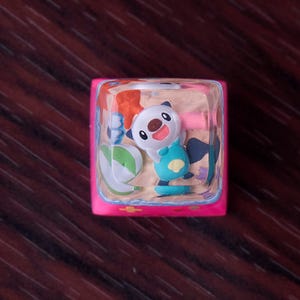 May include: A square mechanical keyboard keycap with a bright pink base and a clear resin top. Inside the resin, a miniature Oshawott character is depicted standing on a sandy beach scene with a green beach ball and a small orange starfish.