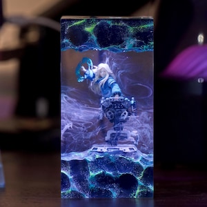 Può includere: Un blocco rettangolare in resina contenente una figura in miniatura con lunghi capelli chiari e un abito blu. La figura è circondata da effetti di fumo bianco vorticoso e accenti verde brillante su uno sfondo blu scuro e strutturato.