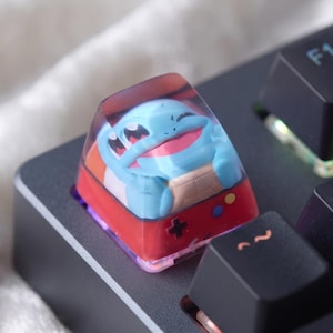 Puede incluir: Una tecla de teclado artesanal personalizada con una parte superior de resina transparente que encierra un personaje de tortuga azul claro con caparazón beige. La base de la tecla es roja e incluye un pad direccional negro y botones circulares pequeños.