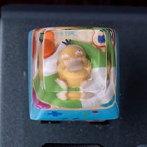 Op de afbeelding: Een vierkante mechanische toetsenbordtoets met een heldere harslaag die een klein geel eendachtig wezen met een platte snavel en drie zwarte haren op het hoofd omsluit, op een lichtblauwe basis met kleine kleurrijke gamecontroller-pictogrammen.