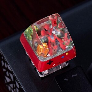 May include: A custom mechanical keyboard keycap featuring a clear resin top encasing a miniature red insectoid figure, green foliage, and rocky textures. The base is solid red with a black plus sign, a blue dot, and a yellow dot printed on the front.