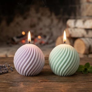May include: Two spherical candles with a spiraled, ribbed texture. One candle is light purple and the other is light green. Both candles are lit, each featuring a single flame at the top, emitting a small wisp of smoke from the burning wicks.