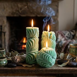 May include: Three sage green candles with intricate swirling textures. One tall cylindrical candle, one medium textured pillar, and one small spherical candle are lit, with a thin trail of smoke rising from the center pillar candle.