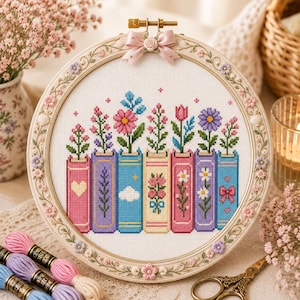Puede incluir: Un bastidor de bordado circular con un patrón de punto de cruz de siete libros coloridos dispuestos verticalmente, cada uno coronado por una flor diferente. El marco está decorado con pequeñas flores esculpidas rosas y moradas y un lazo rosa.