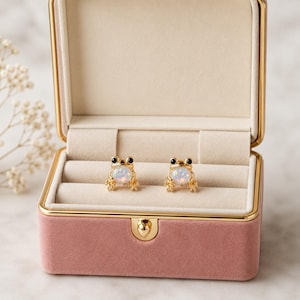 May include: A pair of gold-toned stud earrings shaped like frogs, each featuring a round, iridescent white opal-style stone as the belly and small black beads for the eyes. The earrings are displayed inside a rectangular, pink velvet jewelry box.