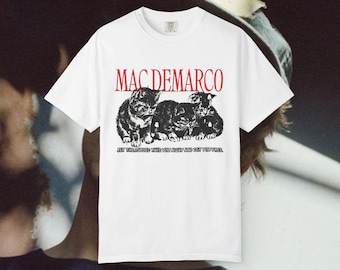Op de afbeelding: Een wit T-shirt met korte mouwen en een zwart-witte afbeelding van drie kittens. Boven de kittens staat de tekst MAC DEMARCO in het rood gedrukt. Onder de kittens staat de tekst LET THE MUSIC TAKE YOU AWAY AND SET YOU FREE in het zwart.