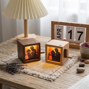 May include: Two square wooden music boxes with internal illuminated photo displays showing silhouettes of a couple. Each box features a side crank handle and a warm light source. The boxes are crafted from dark and light wood, respectively.
