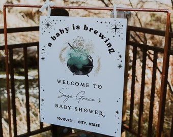 May include: A white sign with black text and a black and green illustration of a cauldron with steam coming out of it. The text reads "a baby is brewing" and "Welcome to Sage Grace's Baby Shower 10-15-22 CITY, STATE".