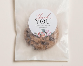 May include: A white favor bag with a round sticker that says "Thank You" in red script with a black cross and the text "Sage's Baby Shower 10.10.2023" in black. The sticker has a watercolor floral design with pink and red flowers.