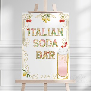 Puede incluir: Un cartel blanco con letras doradas y detalles florales que dice "Italian Soda Bar". El cartel est&aacute; en un caballete y presenta una soda italiana rosa y blanca con una cereza en la parte superior.