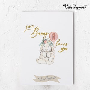 May include: A watercolor illustration of a white bunny wearing a floral crown and holding a pink balloon with the number "1" on it. The text "some bunny loves you" is written in gold script.