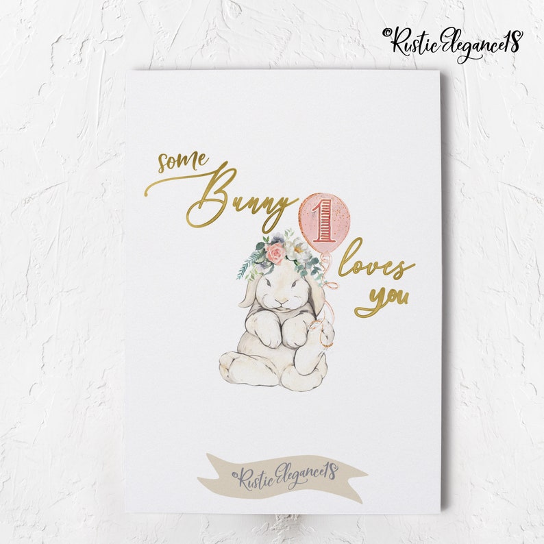 May include: A watercolor illustration of a white bunny wearing a floral crown and holding a pink balloon with the number "1" on it. The text "some bunny loves you" is written in gold script.