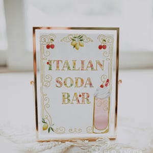 May include: A gold framed sign with a white background and gold lettering that reads "ITALIAN SODA BAR". The sign is decorated with lemons, cherries, and a glass of pink soda with whipped cream.
