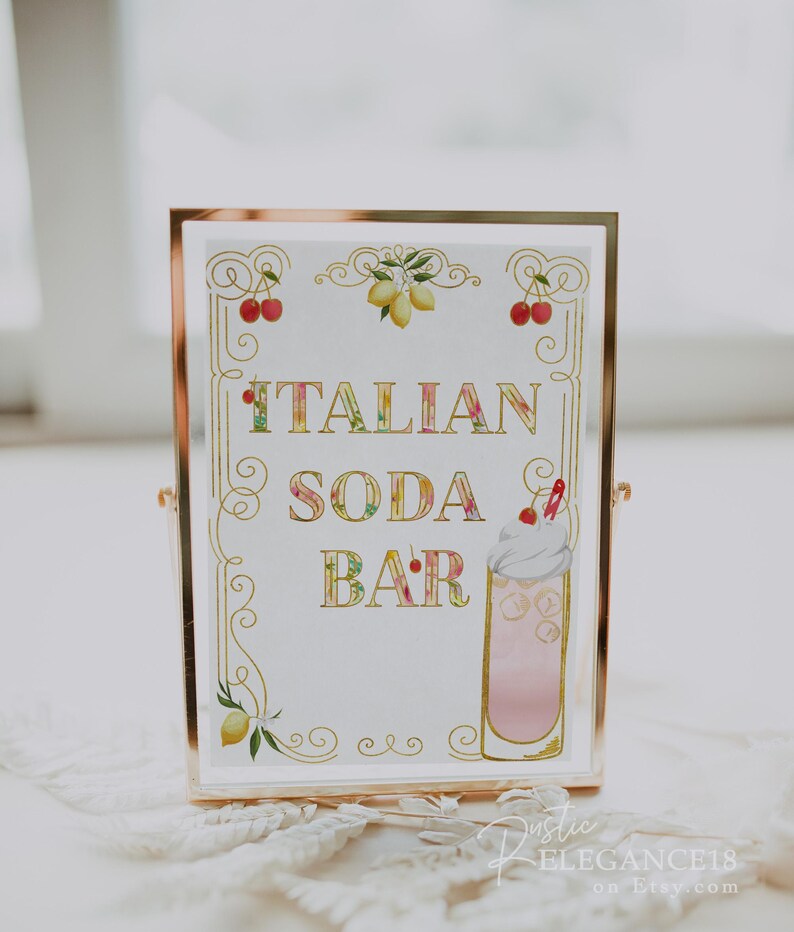 May include: A gold framed sign with a white background and gold lettering that reads "ITALIAN SODA BAR". The sign is decorated with lemons, cherries, and a glass of pink soda with whipped cream.