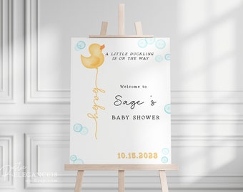 May include: A white welcome sign with a yellow rubber ducky balloon, blue bubbles, and the text "A little duckling is on the way. Welcome to Sage's Baby Shower 10.15.2028".