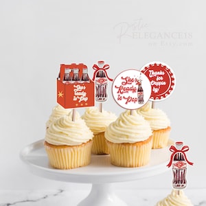 Puede incluir: Cupcakes con glaseado blanco y adornos decorativos con un diseño de botella de refresco vintage. Los adornos incluyen un paquete de seis botellas, botellas individuales con lazos rojos y diseños circulares con texto. Los cupcakes están en un soporte blanco.