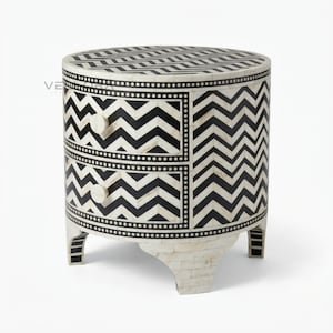 May include: A round bedside table featuring two drawers with a black and white chevron pattern. The surface and drawer fronts are decorated with a geometric zigzag design, accented by a dotted border. It stands on four short, curved legs.
