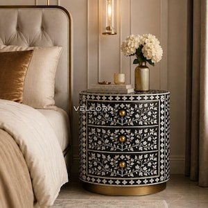 May include: A cylindrical three-drawer nightstand featuring a black finish with intricate white floral inlay patterns. The piece includes three round gold-toned knobs, a solid gold-toned base, and the text VELLORA printed on the front surface.