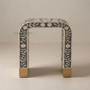 May include: A U-shaped console table featuring a dark gray surface inlaid with white floral and vine patterns. The base of the table is finished with solid gold-toned metal feet. The text VELDORA is visible on the left side of the table frame.