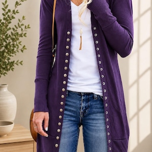May include: A long-sleeved, deep purple cardigan featuring a vertical row of metallic snap buttons along the front opening. The garment has ribbed cuffs and a ribbed hem, worn over a plain white V-neck top and blue denim jeans with a brown bag strap.