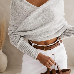 Può includere: Un maglione grigio chiaro a maniche lunghe con scollo a V incrociato e polsini a coste, abbinato a una gonna bianca con bottoni frontali, una cintura in pelle marrone con fibbia dorata e una borsa strutturata marrone con motivo traforato.