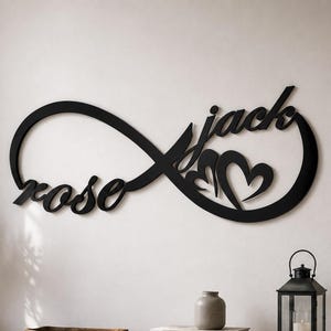 Può includere: Un'insegna da parete nera a forma di simbolo dell'infinito. Il testo in corsivo "rose" è integrato nell'anello sinistro, mentre il testo "jack" è integrato nell'anello destro, accompagnato da due forme a cuore all'interno del design.