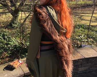 May include: A woman wearing a green hooded tunic with a brown fur trim and a brown leather belt. She has long red hair and is wearing pointed elf ears.