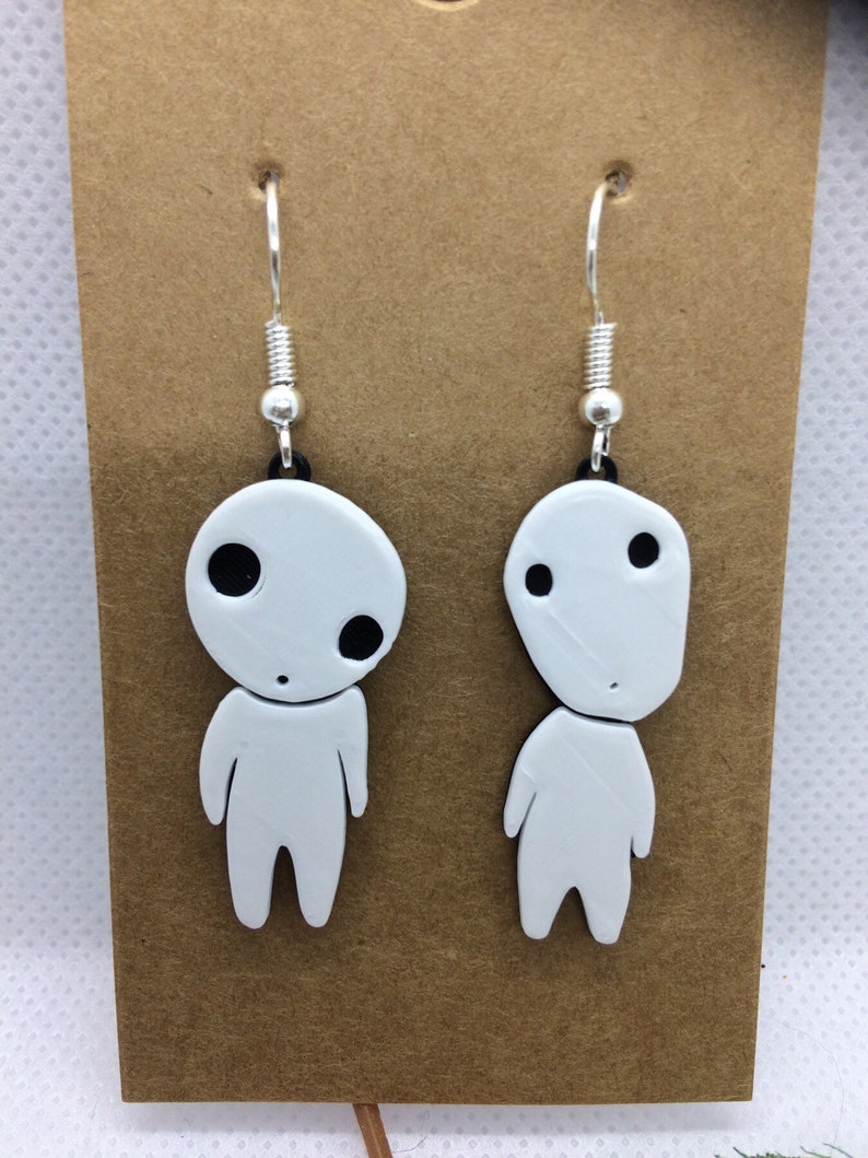 May include: A pair of white dangle earrings featuring a cartoon character with black eyes and a simple smile. The earrings are made of polymer clay and have silver hooks.