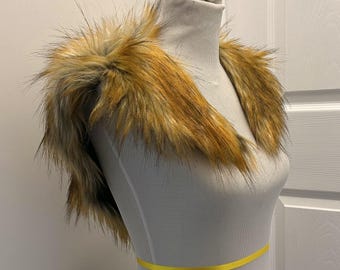 May include: A faux fur collar in shades of brown and gold, displayed on a mannequin. The collar has a fluffy texture and is designed to be worn around the shoulders. A yellow ribbon is wrapped around the mannequin's waist.